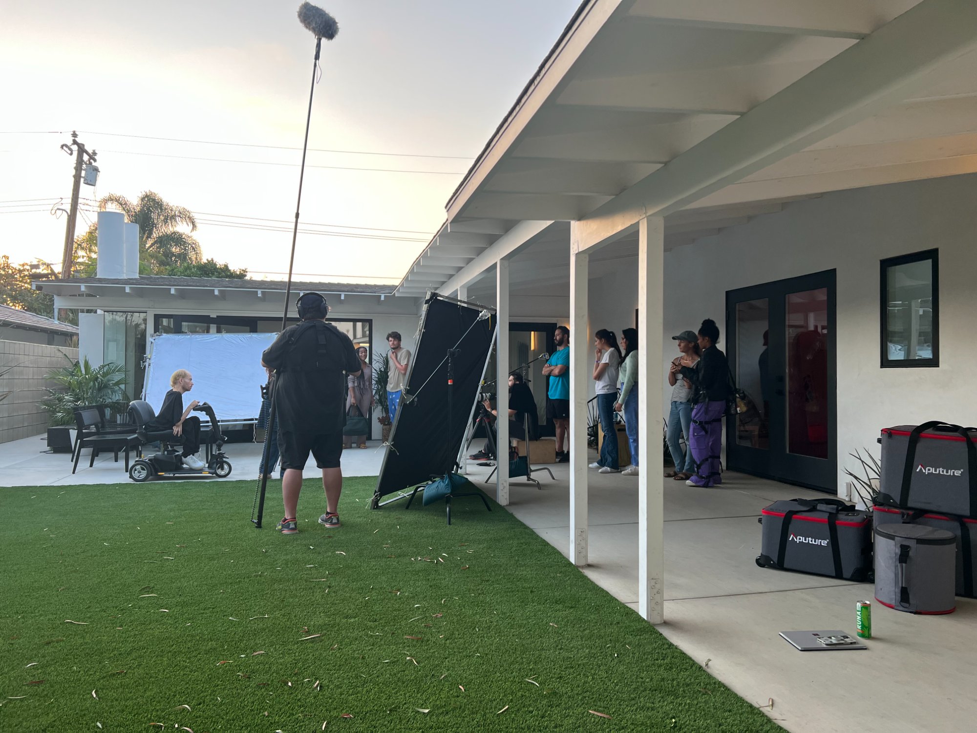Backyard patio scene — bounce board, floppy, boom op, Aputure bags lined up, crew watching between takes