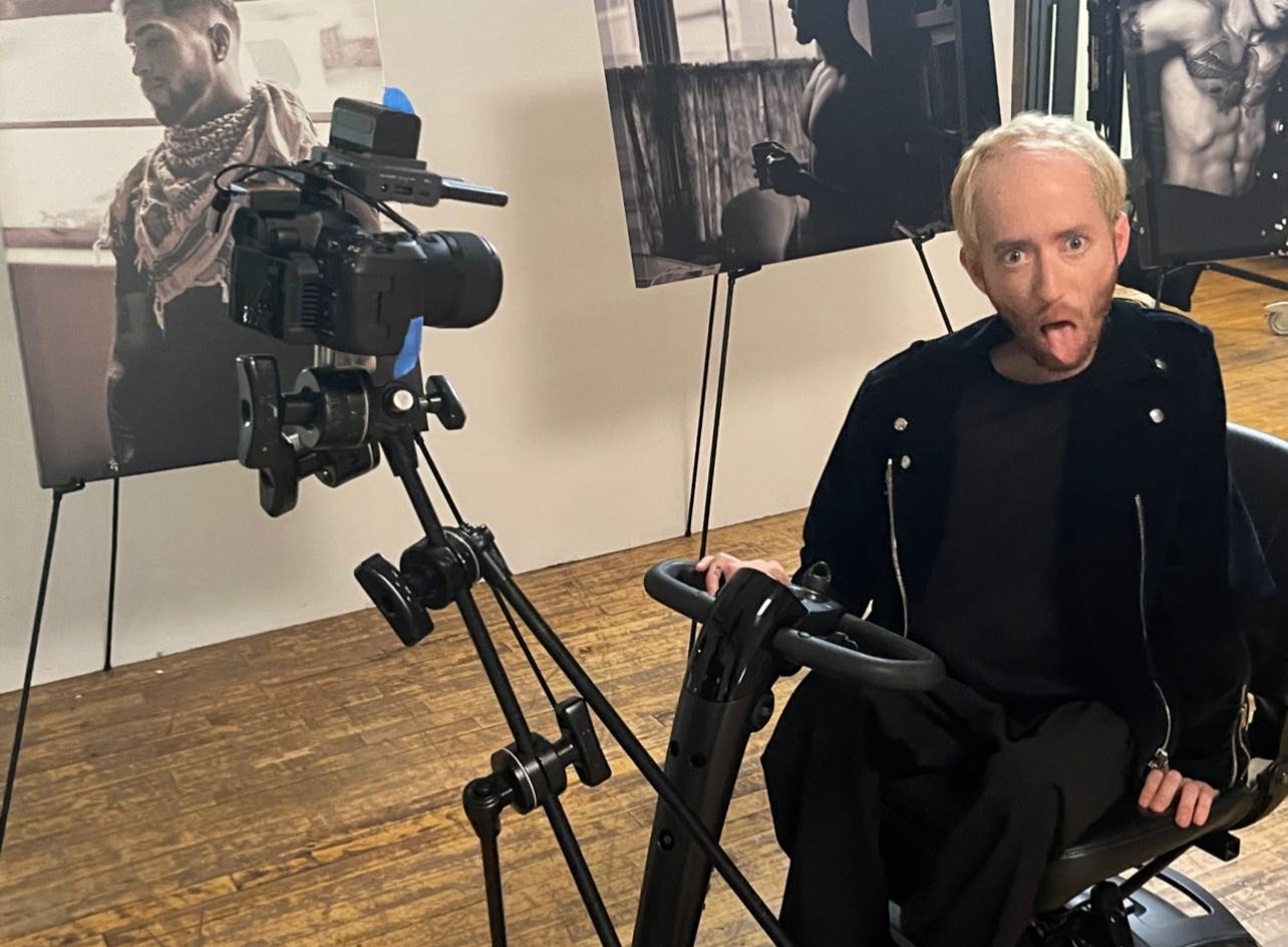 Danny making a face at the camera between takes — photography prints visible in the background